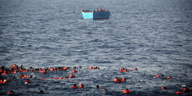 Szene auf offenem Meer, im Hintergrund ein blaues Holzboot, im Vordergrund viele Menschen in Schwimmwesten im Wasser Szene auf offenem Meer, im Hintergrund ein blaues Holzboot, im Vordergrund viele Menschen in Schwimmwesten im Wasser