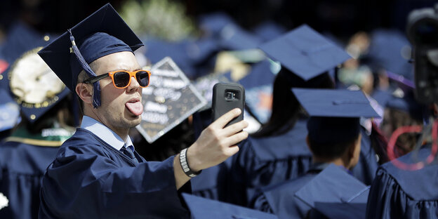 Ein Universitäts-Absolvent in Talar trägt eine Sonnenbrille, streckt die Zunge raus und macht eine Selfie mit seinem Handy Ein Universitäts-Absolvent in Talar trägt eine Sonnenbrille, streckt die Zunge raus und macht eine Selfie mit seinem Handy