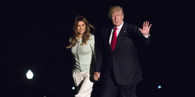 Trump schreitet, seine Frau Melania an der Hand, forsch durch die Dunkelheit und winkt Trump schreitet, seine Frau Melania an der Hand, forsch durch die Dunkelheit und winkt