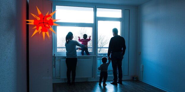 Eine Familie steht in der Dämmerung am Fenster ihrer unbeleuchteten Wohnung und schaut in die Ferne Eine Familie steht in der Dämmerung am Fenster ihrer unbeleuchteten Wohnung und schaut in die Ferne