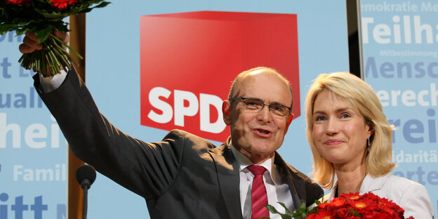 Ein Mann und eine Frau nebeneinander, beide haben jeweils einen roten Blumenstrauß in der Hand, der Mann hält seinen in die Luft Ein Mann und eine Frau nebeneinander, beide haben jeweils einen roten Blumenstrauß in der Hand, der Mann hält seinen in die Luft