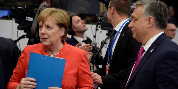 Merkel, in rotem Jackett, hält eine blaue Mappe fest in ihren Händen und guckt nach links, rechts von ihr steht Orban und guckt zu ihr Merkel, in rotem Jackett, hält eine blaue Mappe fest in ihren Händen und guckt nach links, rechts von ihr steht Orban und guckt zu ihr