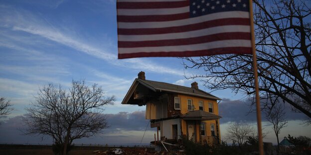 Ein zerstörtes Haus im Hintergrund vor einem dämmernden Himmel, im Vordergrund eine wehenede US-Flagge Ein zerstörtes Haus im Hintergrund vor einem dämmernden Himmel, im Vordergrund eine wehenede US-Flagge