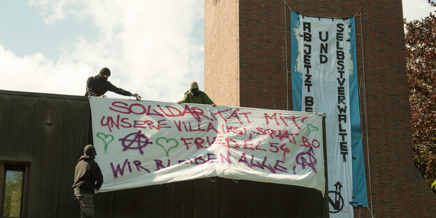 Maskierte hängen ein Spruchbanner auf einem Kirchendach auf.