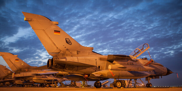Drei Bundeswehr-Tornados stehen im rötlichen Lich von Scheinwerfern auf einem Rollfeld vor dunkel bewölktem Himmel Drei Bundeswehr-Tornados stehen im rötlichen Lich von Scheinwerfern auf einem Rollfeld vor dunkel bewölktem Himmel