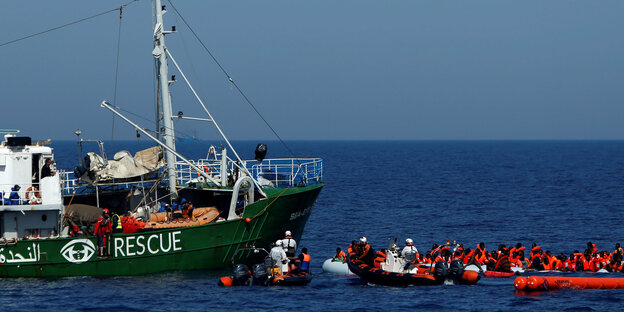 Ein Rettungsschiff mit der Aufschrift "Rescue" nimmt Geflüchtete in orangenen Rettungswesten auf, die vor dem Schiff in Rettungsbooten warten Ein Rettungsschiff mit der Aufschrift "Rescue" nimmt Geflüchtete in orangenen Rettungswesten auf, die vor dem Schiff in Rettungsbooten warten