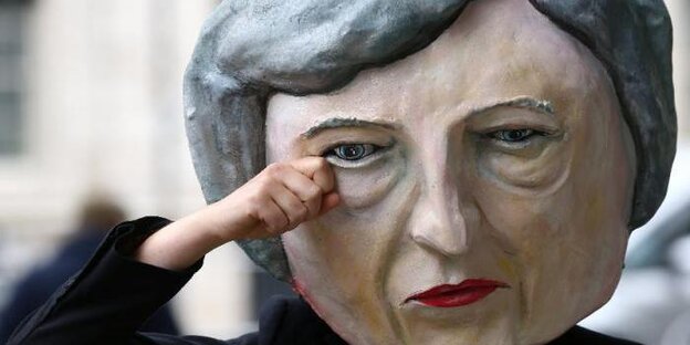 Eine Hand fässt an eine Pappmaske. Sie zeigt das Gesicht von Theresa May in Übergröße Eine Hand fässt an eine Pappmaske. Sie zeigt das Gesicht von Theresa May in Übergröße