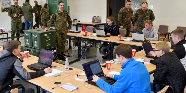 Ein paar Jugendliche sitzen mit Laptops an einem Tisch, davor stehen Menschen in Bundeswehr-Uniform Ein paar Jugendliche sitzen mit Laptops an einem Tisch, davor stehen Menschen in Bundeswehr-Uniform