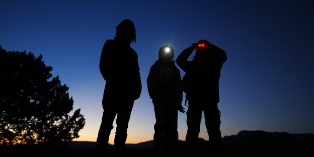 Drei Personen stehen im schwindenden Licht auf einem Hügel und blicken durch Nachtsichtgeräte Drei Personen stehen im schwindenden Licht auf einem Hügel und blicken durch Nachtsichtgeräte