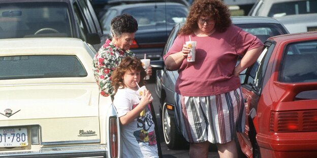 Zwei Frauen und ein Kind stehen zwischen Autos und trinken etwas Zwei Frauen und ein Kind stehen zwischen Autos und trinken etwas