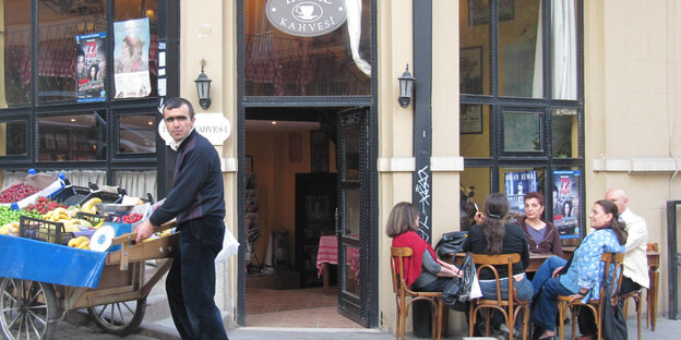 Menschen sitzen gemütlich vor einem Cafe, ein Mann steht an einem Obststand Menschen sitzen gemütlich vor einem Cafe, ein Mann steht an einem Obststand