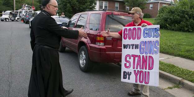 Ein Priester streckt den Arm einem Mann mit Plakat zum Handschlag aus, auf dem steht: "Good guys with guns stand by you" Ein Priester streckt den Arm einem Mann mit Plakat zum Handschlag aus, auf dem steht: "Good guys with guns stand by you"
