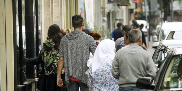 Menschen laufen zwischen Häuserfronten und geparkten Autos auf einem Gehweg Menschen laufen zwischen Häuserfronten und geparkten Autos auf einem Gehweg