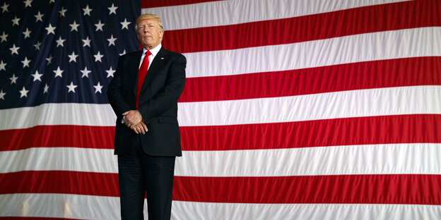 Ein Mann im Anzug steht vor einer riesengroßen US-Flagge Ein Mann im Anzug steht vor einer riesengroßen US-Flagge