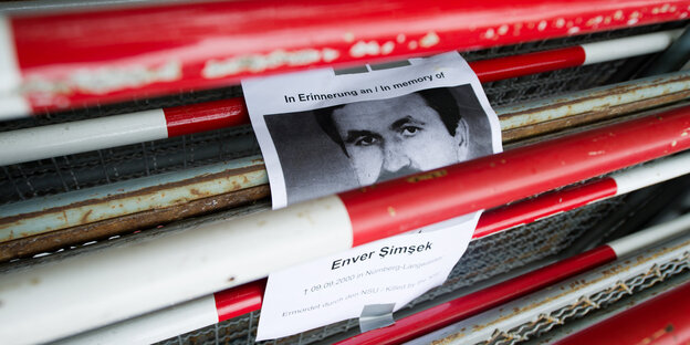 Zwischen rot-weißen Absperrgittern hängt ein Papier mit der Aufschrift „In Erinnerung an / In memory of - Enver Simsek - 09.09.2000 - Ermordet durch den NSU / Killed by the NSU" Zwischen rot-weißen Absperrgittern hängt ein Papier mit der Aufschrift „In Erinnerung an / In memory of - Enver Simsek - 09.09.2000 - Ermordet durch den NSU / Killed by the NSU"