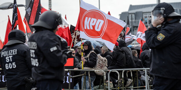 Ein Demonstrant in einer Menschenmenge trägt eine NPD-Fahne, vor ihm stehen Polizisten in Vollmontur