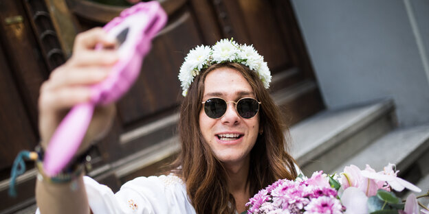 Ein junger Mann mit Sonnenbrille, langen Haaren und Blumenkranz filmt sich selbst mit einem pinken Handy Ein junger Mann mit Sonnenbrille, langen Haaren und Blumenkranz filmt sich selbst mit einem pinken Handy