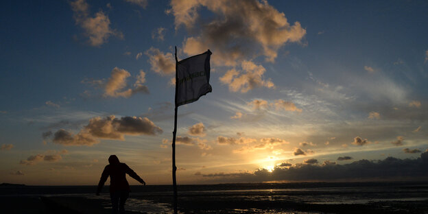 Eine Fahne weht im Wind, dahintertaucht die Sonne ins Meer, ein Mann schreitet durch die Dämmerung Eine Fahne weht im Wind, dahintertaucht die Sonne ins Meer, ein Mann schreitet durch die Dämmerung