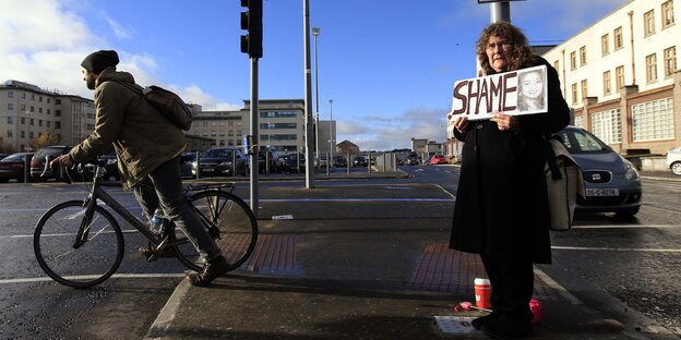 Eine Frau hält ein Schild in die Höhe, auf dem „Shame“ (Schande) steht, im Hintergrund ein Fahrradfahrer Eine Frau hält ein Schild in die Höhe, auf dem „Shame“ (Schande) steht, im Hintergrund ein Fahrradfahrer