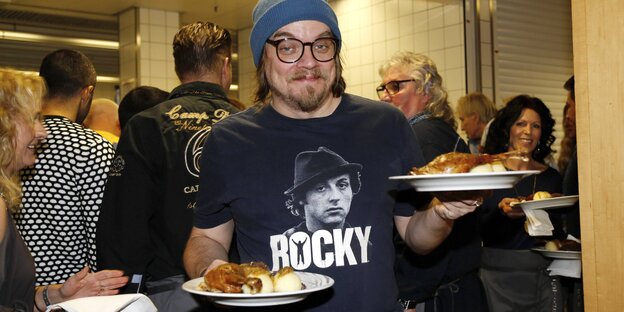 Mann mit Wollmütze, dicker Brille und "Rocky"-T-Shirt schneidet Grimasse und balanciert zwei Teller mit Essen Mann mit Wollmütze, dicker Brille und "Rocky"-T-Shirt schneidet Grimasse und balanciert zwei Teller mit Essen
