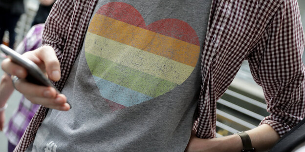 Ein Mann mit einem regenbogenfarbenen Herz auf dem T-Shirt tippt mit einer Hand auf seinem Handy Ein Mann mit einem regenbogenfarbenen Herz auf dem T-Shirt tippt mit einer Hand auf seinem Handy