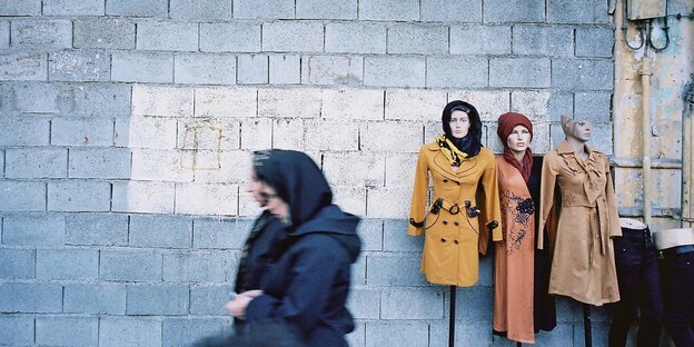 Zwei Frauen in dunklen Mänteln und Kopftüchern gehen an Kleiderständern mit bunten Outfits vorbei Zwei Frauen in dunklen Mänteln und Kopftüchern gehen an Kleiderständern mit bunten Outfits vorbei