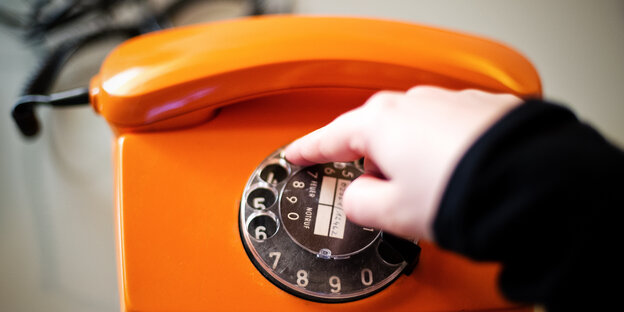 Eine Hand wählt an der Drehscheibe eines alten, orangefarbenen Telefons eine Nummer Eine Hand wählt an der Drehscheibe eines alten, orangefarbenen Telefons eine Nummer