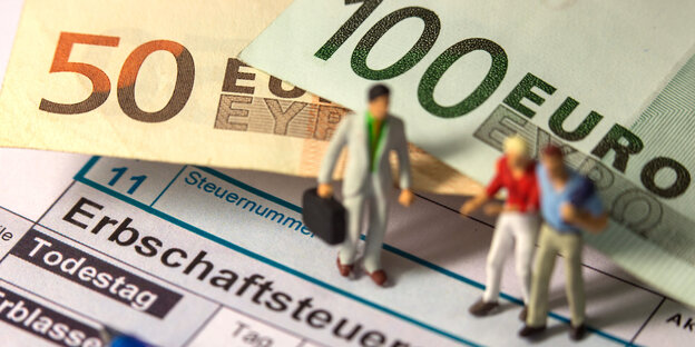 Man sieht ein Formular zur Erbschaftssteuererklärung, einen 50- und einen 100-Euroschein und zwei Modellfiguren Man sieht ein Formular zur Erbschaftssteuererklärung, einen 50- und einen 100-Euroschein und zwei Modellfiguren