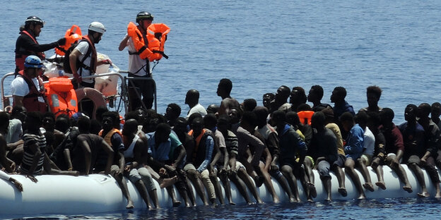 Migranten sitzen in einem überfüllten Schlauchboot auf dem Mittelmeer. Helfer von Hilfsorganisationen nähern sich ihnen und verteilen Schwimmwesten Migranten sitzen in einem überfüllten Schlauchboot auf dem Mittelmeer. Helfer von Hilfsorganisationen nähern sich ihnen und verteilen Schwimmwesten