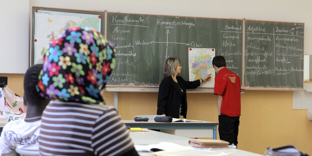 Ein Schüler und eine Lehrerin stehen an der Tafel, ein Mädchen mit Kopftuch schaut ihnen zu Ein Schüler und eine Lehrerin stehen an der Tafel, ein Mädchen mit Kopftuch schaut ihnen zu