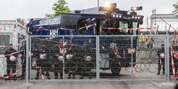 ein Wasserwerfer vor dem Polizisten stehen hinter einem Zaun mit Nato-Draht ein Wasserwerfer vor dem Polizisten stehen hinter einem Zaun mit Nato-Draht