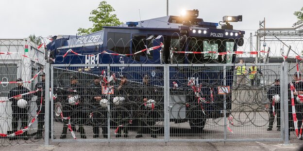 Polizisten und ein Wasserwerfer stehen hinter einem Zaun