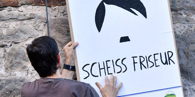 Mann hängt Plakat mit Hitler-Frisur und -Bart an eine Wand. Aufschrift: Scheiss Friseur Mann hängt Plakat mit Hitler-Frisur und -Bart an eine Wand. Aufschrift: Scheiss Friseur