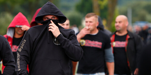 Ein Mann zieht sich seinen Kapuzenpulli ins Gesicht. Hinter ihm laufen weitere Männer Ein Mann zieht sich seinen Kapuzenpulli ins Gesicht. Hinter ihm laufen weitere Männer