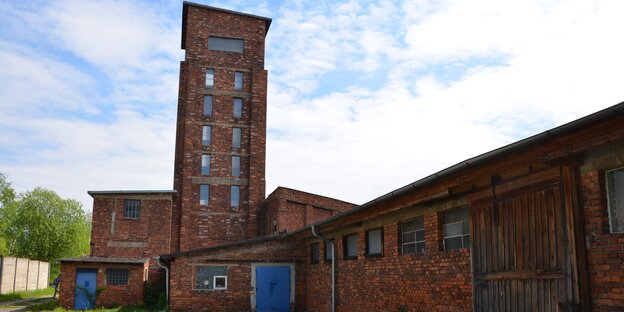 ein langgestrecktes Gebäude, das von einem hohen Turm mit Fenstern überragt wird. Die Bauten sind aus rotem Ziegel ein langgestrecktes Gebäude, das von einem hohen Turm mit Fenstern überragt wird. Die Bauten sind aus rotem Ziegel