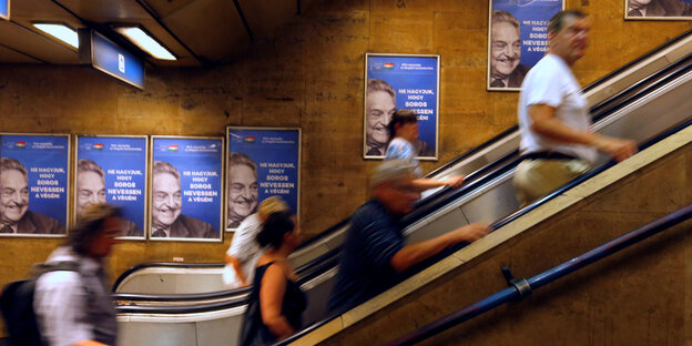 Personen auf einer Rolltreppe, im Hintergrund Poster mit dem Gesicht von George Soros Personen auf einer Rolltreppe, im Hintergrund Poster mit dem Gesicht von George Soros