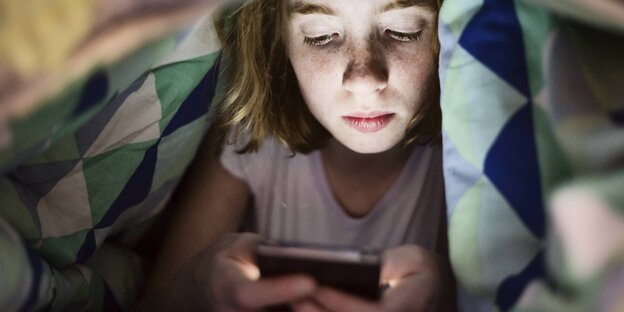 Ein Mädchen tippt unter der Bettdecke auf dem Smartphone rum Ein Mädchen tippt unter der Bettdecke auf dem Smartphone rum
