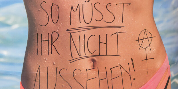 Auf einem Werbeplakat, das eine Frau im Bikini zeigt, steht per Hand geschrieben: "SO MÜSST IHR NICHT AUSSEHEN!" Auf einem Werbeplakat, das eine Frau im Bikini zeigt, steht per Hand geschrieben: "SO MÜSST IHR NICHT AUSSEHEN!"