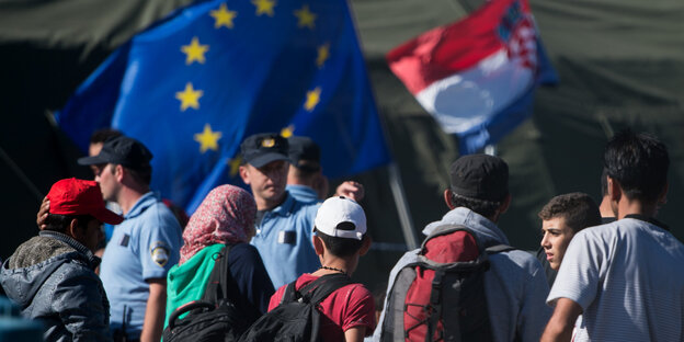 Menschen mit Rucksäcken stehen vor Polizisten und Fahnen von Europa und Kroatien Menschen mit Rucksäcken stehen vor Polizisten und Fahnen von Europa und Kroatien