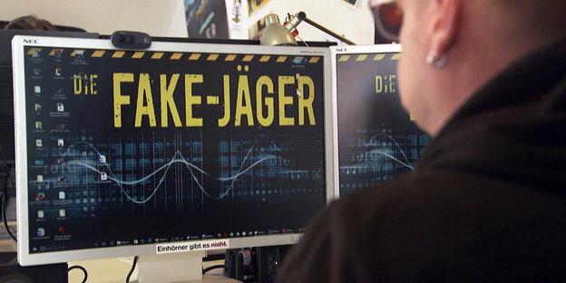 Ein Mann sitzt vor einem großen Computer-Bildschirm, auf dem in gelber Schrift "Fake-Jäger" steht Ein Mann sitzt vor einem großen Computer-Bildschirm, auf dem in gelber Schrift "Fake-Jäger" steht