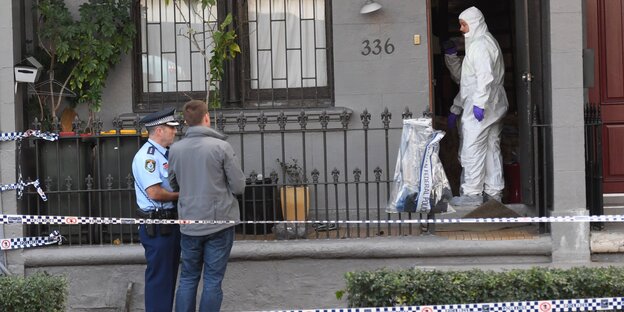 Ein Polizist, ein Mann in einem weißen Ganzkörperanzug und eine weiterer Mann stehen vor einem Haus Ein Polizist, ein Mann in einem weißen Ganzkörperanzug und eine weiterer Mann stehen vor einem Haus