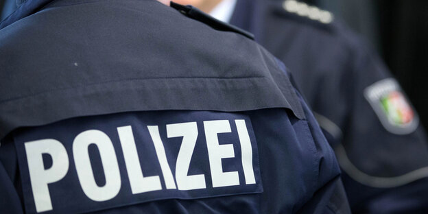 Der Rücken eines Polizisten, darauf der Schriftzug "POLIZEI". Im Hintergrund ein weiterer Polizist. Der Rücken eines Polizisten, darauf der Schriftzug "POLIZEI". Im Hintergrund ein weiterer Polizist.