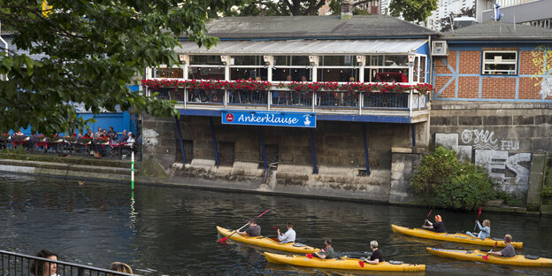 Landwehrkanal an der Kottbusser Brücke, auf dem Kanal sind vier gelbe Kanus, im Hintergrund die Kneipe "Ankerklause", prominent zu sehen der Balkon.