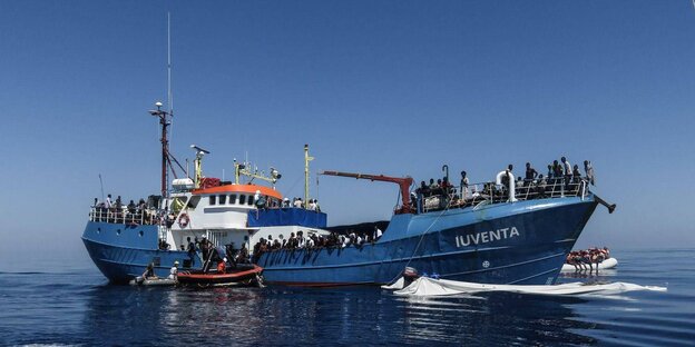 ein großes Schiff mit zahlreichen Personen an Bord, davor und dahinter mehrere kleinere Boote