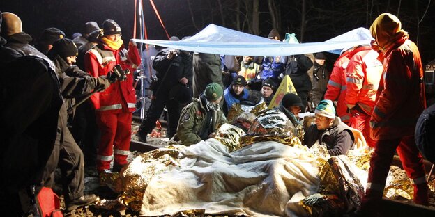 Es ist Nacht, Menschen liegen zugedeckt und mit Mützen auf dem Boden, um sie herum viele andere Menschen