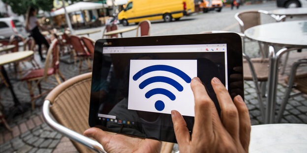 Zwei Hände halten in einem Café, draußen, ein Tablet in die Höhe. Auf dem Bildschirm ist ein Symbol WLAN-Symbol zu sehen. Zwei Hände halten in einem Café, draußen, ein Tablet in die Höhe. Auf dem Bildschirm ist ein Symbol WLAN-Symbol zu sehen.