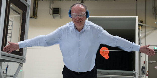Ein Mann in weißem Hemd hat eine Schutzbrille und Ohrenschützer auf und streckt beide Arme zur Seite Ein Mann in weißem Hemd hat eine Schutzbrille und Ohrenschützer auf und streckt beide Arme zur Seite