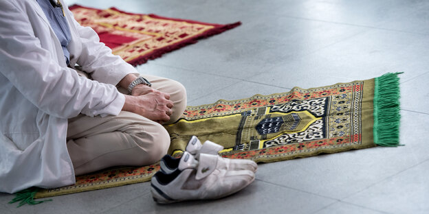 Eine Person kniet auf einem Gebetsteppich und hat ihre Turnschuhe neben ihm abgestellt. Eine Person kniet auf einem Gebetsteppich und hat ihre Turnschuhe neben ihm abgestellt.