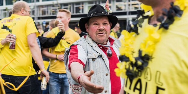 Männer in Fußballtrikots stehen auf einem Platz. In der Mitte steht ein Mann mit Hut, der einem anderen seine Hand entgegenstreckt. Männer in Fußballtrikots stehen auf einem Platz. In der Mitte steht ein Mann mit Hut, der einem anderen seine Hand entgegenstreckt.