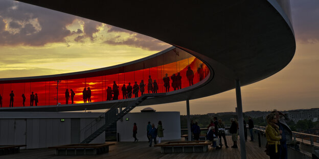 Eine geschwungene, verglaste Brücke mit rot getönten Scheiben vor Sonnenuntergang mit Menschen Eine geschwungene, verglaste Brücke mit rot getönten Scheiben vor Sonnenuntergang mit Menschen
